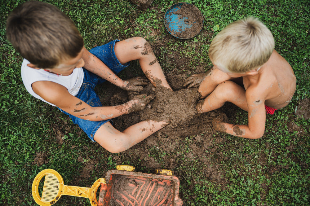Two young boys playing joyfully in a mud puddle on green grass, their hands and legs covered in dirt, embodying carefree, nature-based childhood.