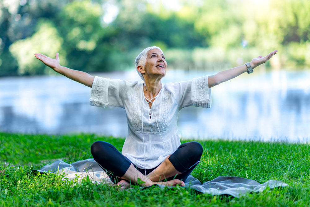 Joyful senior woman practicing mindfulness outdoors, sitting cross-legged on grass by a lake with arms raised, symbolizing presence, peace, and well-being.