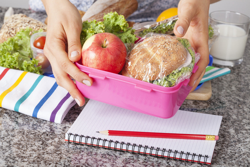 Pink lunchbox filled with fresh apple, whole grain sandwich, on table with notebook and pencil.
