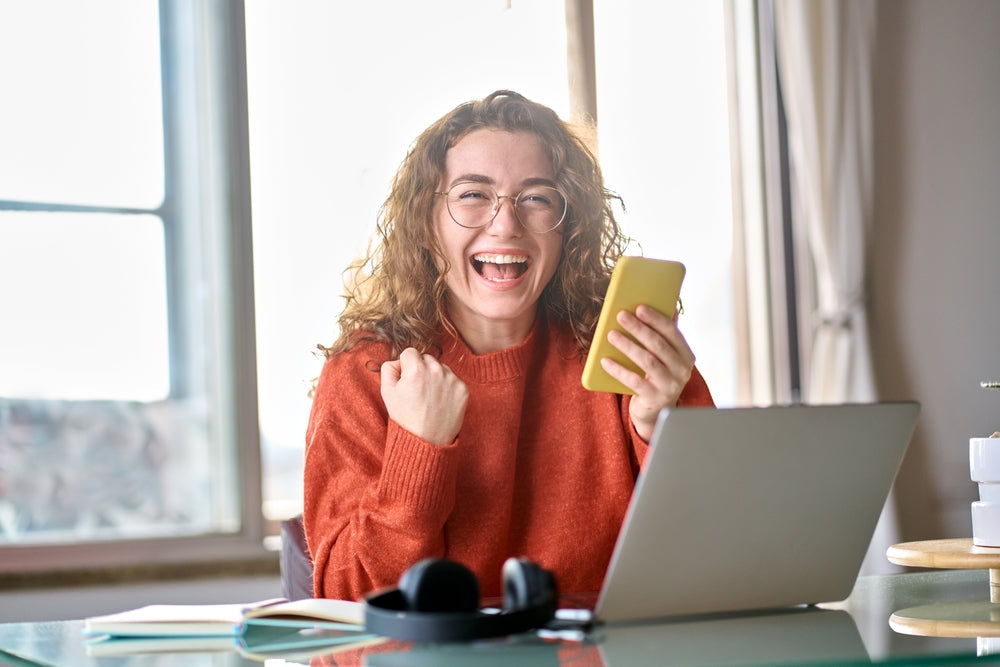 Happy woman working online from home, smiling and celebrating success with a smartphone and laptop.