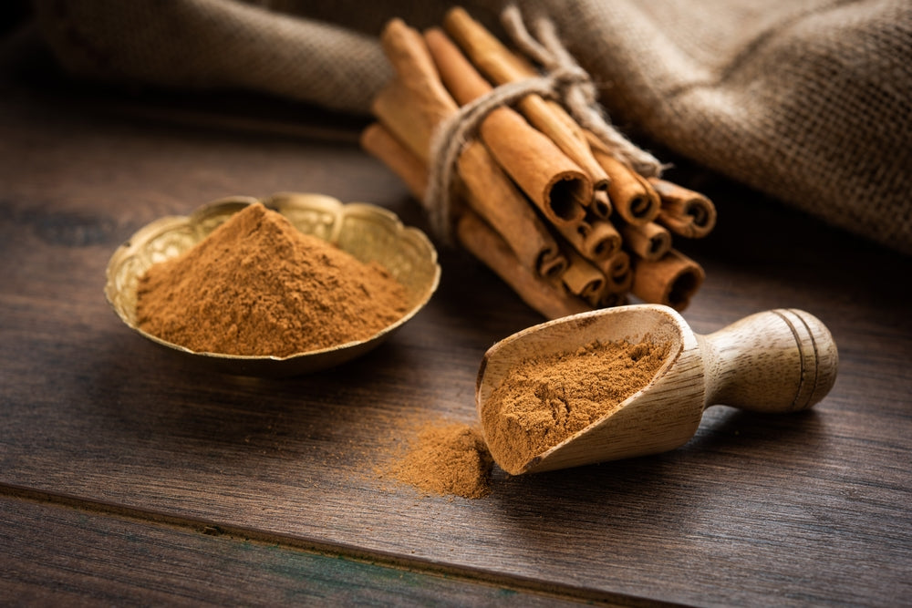Ground cinnamon in a wooden scoop and golden bowl beside cinnamon sticks tied with twine on a rustic wooden table.