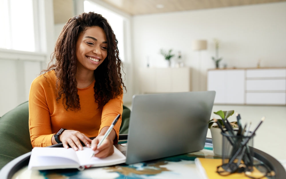 Smiling woman working from home on her laptop, taking notes while learning how to build a successful online business.